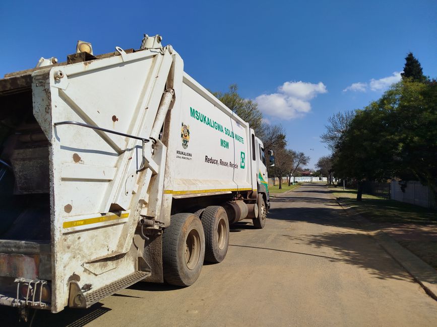 A white waste collection truck is parked on a residential street, positioned to the left side of the image. The truck has a large, rectangular rear compartment with visible signs of dirt and rust on i