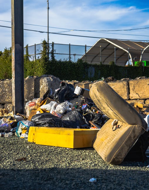 A worker wearing a high-visibility yellow and orange safety vest is performing waste collection activities in front of a large, red refuse collection truck parked on the side of a street. The truck's 