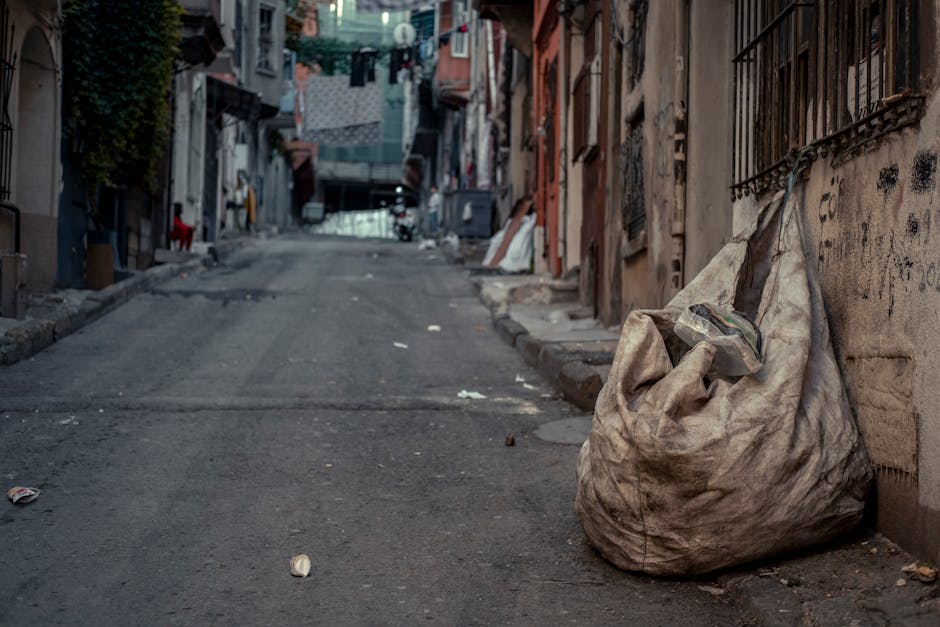 Two large black plastic garbage bags, filled and tied at the top, are situated on the edge of a paved street beside a curb. The bags contain household waste, possibly including paper and plastic packa